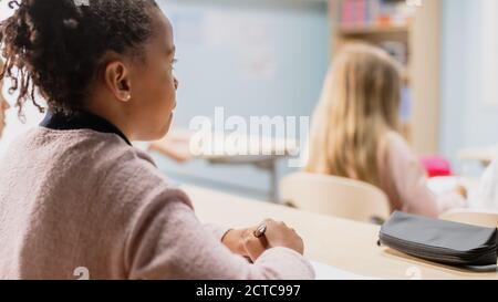 En classe d'école élémentaire : vue sur l'épaule d'une fille noire brillante à l'écoute d'un enseignant. Salle de classe junior avec groupe varié de lumineux Banque D'Images