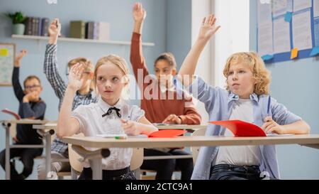 Salle de classe élémentaire de divers enfants à l'écoute de l'enseignant donner une leçon. Tout le monde soulève la main en sachant la bonne réponse. Jeunes enfants à l'école Banque D'Images