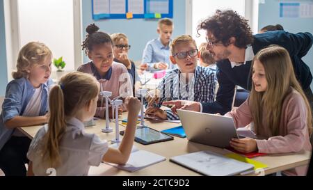 École primaire salle de classe: Enseignant enthousiaste tenant un ordinateur de tablette explique à un brillant jeunes enfants comment les éoliennes fonctionnent. Apprentissage des enfants Banque D'Images