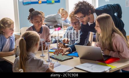 École primaire salle de classe: Enseignant enthousiaste tenant un ordinateur de tablette explique à un brillant jeunes enfants comment les éoliennes fonctionnent. Apprentissage des enfants Banque D'Images