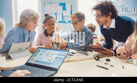 École primaire robotique salle de classe: Groupe diversifié d'enfants brillants avec enseignant enthousiaste de construction et de programmation robot. Apprentissage des enfants Banque D'Images