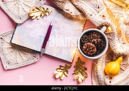 Des cônes de pin dans une tasse en émail gris, un carnet avec un stylo, des feuilles dorées, une guirlande lumineuse et un motif écossais tricoté sur fond rose. Plat, haut vi Banque D'Images