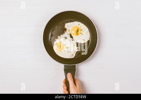 Femme tenant main poêle avec oeufs cuits frits sur table en bois blanc, vue de dessus. Banque D'Images