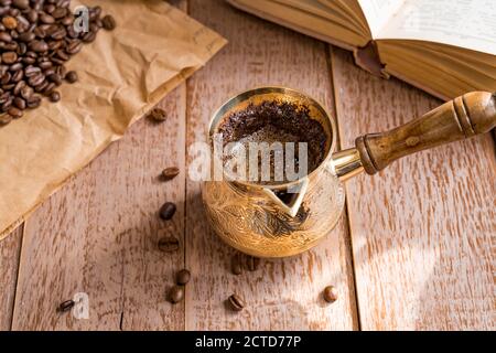 café fraîchement préparé dans une cafetière turque traditionnelle, livre ouvert et grains de café sur une table en bois. Banque D'Images