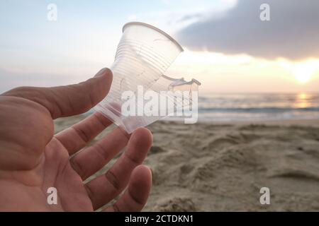 Homme main gros plan avec verre plastique isposable jeté sur sable côte de mer, pollution Banque D'Images