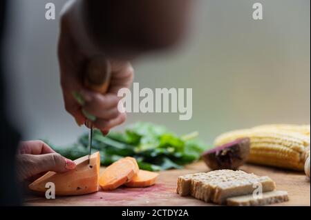 Vue à angle bas d'une femme coupant de la patate douce sur une planche à découper pleine de légumes et de protéines de tempeh. Banque D'Images