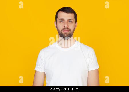 Jeune homme avec une barbe dans un t-shirt blanc avec une expression sérieuse sur son visage. Simple et naturel à regarder dans l'appareil photo. Se trouve sur un arrière-plan jaune isolé. Banque D'Images