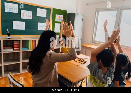 Jeune femme travaillant à l'école avec le bras levé, les élèves mettant leurs mains vers le haut pour répondre à la question, enthousiasme, désireux, plaisir. Professeur d'école asiatique avec des élèves qui élèvent la main Banque D'Images