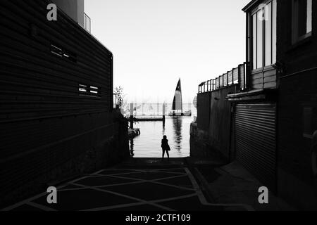 Allée à Cowes Isle of Wight un yacht sombre Mainsail ne passe que par la marina du port pendant qu'une femme montres de la cale de bord de mer Banque D'Images