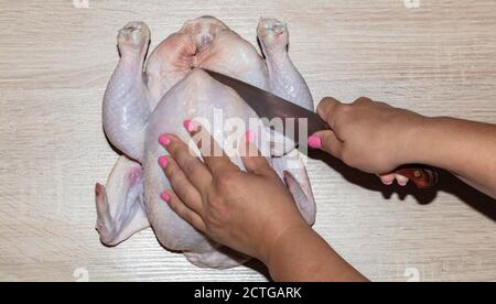 Gros plan d'une femme qui coupe du poulet entier sur une planche en bois. Ongles roses. Banque D'Images