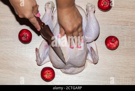 Gros plan d'une femelle coupant du poulet entier sur une planche en bois. Pommes rouges couchés autour du poulet. Ongles roses. Banque D'Images