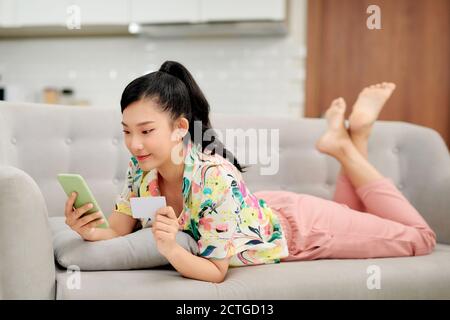 Photo d'une belle jeune femme surpris de brunette sur un canapé à l'intérieur à la maison en utilisant le téléphone portable tenant la carte de crédit. Banque D'Images