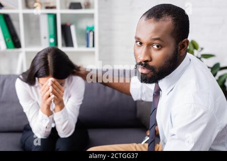 un psychologue afro-américain sérieux regarde la caméra tout en calmant les pleurs femme obscurcissant le visage avec les mains Banque D'Images