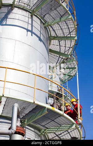 Colonne de distillation grise de l'usine de raffinerie de pétrole sur fond bleu ciel. Société CNPC-AMG. Deux ouvriers d'entretien asiatiques. Banque D'Images