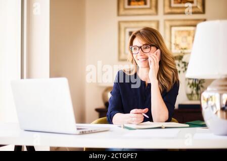 Femme d'affaires confiante assise au bureau et passant un appel téléphonique tout en travaillant à la maison. Bureau à domicile. Banque D'Images