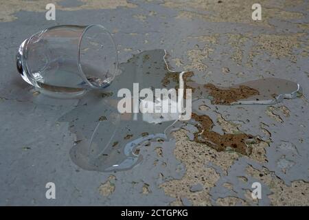 Verre d'eau renversé ou renversé sur la plaque de béton avec écaillage de la vieille peinture. Le liquide renversé forme une flaque irrégulière autour du verre. Banque D'Images