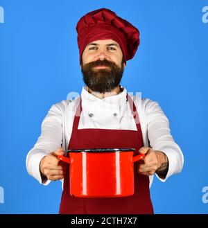 Concept de cuisine professionnelle. Faites cuire avec Happy face dans un chapeau bordeaux et le tablier tient le pot à soupe. Chef avec une casserole ou une casserole rouge. Homme avec barbe tient les ustensiles de cuisine sur fond bleu. Banque D'Images