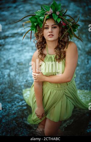 Belle jeune femme dans une robe verte et une couronne dans une forêt ensoleillée. Cérémonie au milieu de l'été ou jour de la Terre. Idée et concept de parfum, contes de fées an Banque D'Images