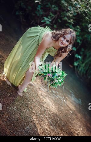 Belle jeune femme dans une robe verte et une couronne dans une forêt ensoleillée. Cérémonie au milieu de l'été ou jour de la Terre. Idée et concept de parfum, contes de fées an Banque D'Images