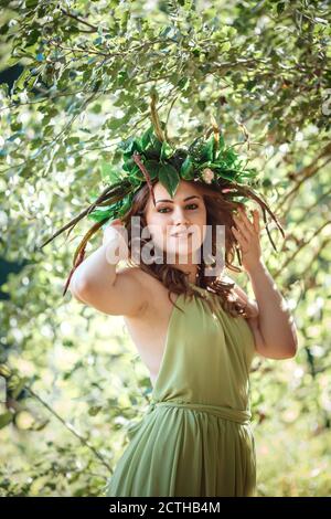 Belle jeune femme dans une robe verte et une couronne dans une forêt ensoleillée. Cérémonie au milieu de l'été ou jour de la Terre. Idée et concept de parfum, contes de fées an Banque D'Images