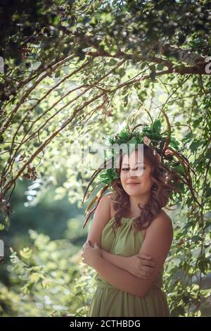 Belle jeune femme dans une robe verte et une couronne dans une forêt ensoleillée. Cérémonie au milieu de l'été ou jour de la Terre. Idée et concept de parfum, contes de fées an Banque D'Images