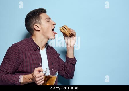 homme gai ivre avec une tasse de bière et un hamburger à la main régime alimentaire style de vie fond bleu Banque D'Images