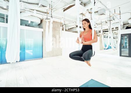 Ariel yoga. Jeune femme belle et calme dans les vêtements de sport assis dans lotus poser dans le hamac blanc suspendu et méditant, gardant les yeux fermés. Bien-être et mode de vie sain, vie en harmonie Banque D'Images