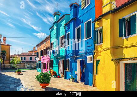 Maisons colorées de l'île de Burano. Bâtiments multicolores et fleurs en pots dans une petite place, ciel bleu en arrière-plan ensoleillé jour d'été, province de Venise, région de Vénétie, Italie du Nord. Carte postale Burano Banque D'Images