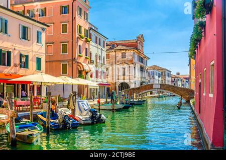 Chioggia paysage urbain avec canal d'eau étroit Vena, bateaux multicolores amarrés, restaurant de rue sur le remblai, vieux bâtiments colorés et pont en brique, ciel bleu en été, région de Vénétie, Italie Banque D'Images