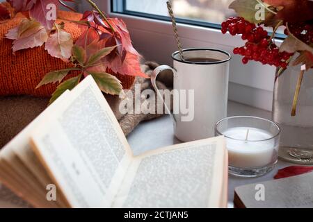Une tasse de café en fer avec une cuillère sur le rebord de la fenêtre au milieu d'un vase avec des branches et des baies de viburnum, un chandail, un livre ouvert et une bougie. Photo pour Banque D'Images