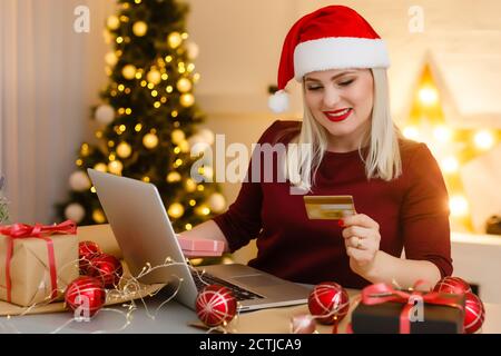 Femme souriante faisant des achats de Noël en ligne comme elle s'assoit dans Devant l'arbre de Noël dans un chapeau de père Noël rouge vérifier les détails de sa carte pour les saisir sur Banque D'Images