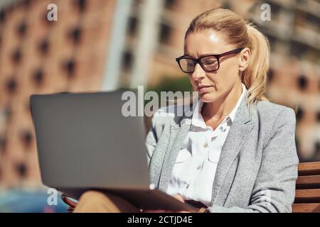 Belle femme d'affaires d'âge moyen travaillant en ligne, utilisant un ordinateur portable tout en étant assise sur le banc à l'extérieur Banque D'Images