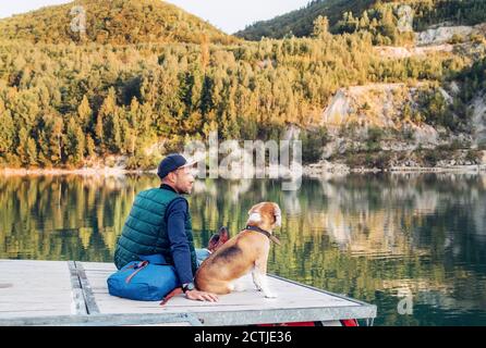 Le propriétaire du chien et son ami le chien beagle sont assis dessus la jetée en bois sur le lac de montagne et de profiter de la paysage pendant leur marche en automne Banque D'Images