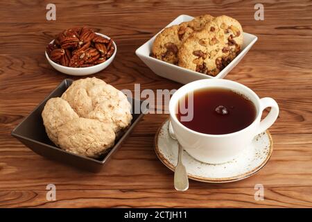 Biscuits aux amandes et aux flocons d'avoine avec noix et raisins secs dans des bols carrés, noix de pécan dans un petit bol, et une tasse de thé noir sur un tabl en bois Banque D'Images