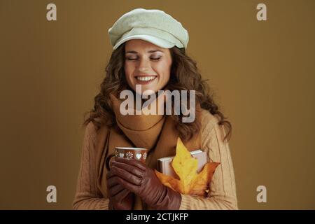 Bonjour novembre. Femme élégante souriante en chandail avec feuille d'érable jaune d'automne, livre, gants en cuir et tasse isolés sur beige. Banque D'Images