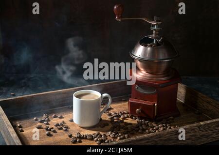 Tasse à café avec boisson chaude à la vapeur, moulin nostalgique et quelques grains torréfiés sur un plateau en bois rustique, fond sombre avec espace de copie, mise au point sélectionnée, Banque D'Images