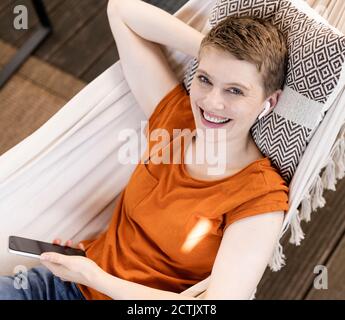 Belle femme souriante utilisant un téléphone portable tout en se reposant sur un hamac dans le porche Banque D'Images