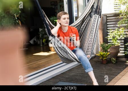 Femme de taille moyenne attentionnés tenant le café tout en étant assise sur une balançoire dans le porche Banque D'Images