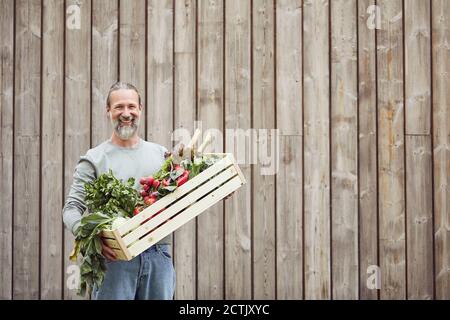 Homme mûr souriant portant une caisse de légumes tout en se tenant contre la maison Banque D'Images