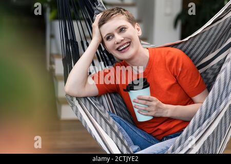 Femme mi-adulte gaie tenant une tasse de café tout en étant assise balancer dans le porche Banque D'Images