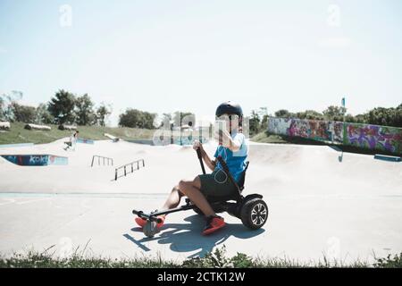 Petit garçon mignon prenant le selfie tout en étant assis sur planekart à parc de skateboard contre le ciel Banque D'Images