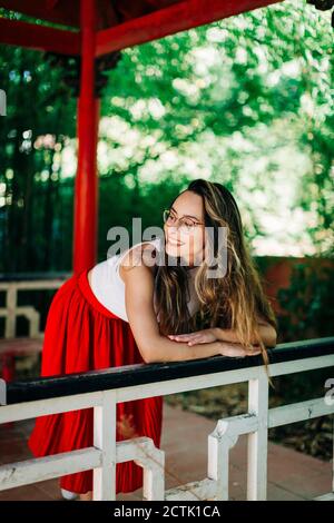 Jeune femme souriante qui regarde loin en se tenant debout près de la main courante passerelle dans le parc Banque D'Images