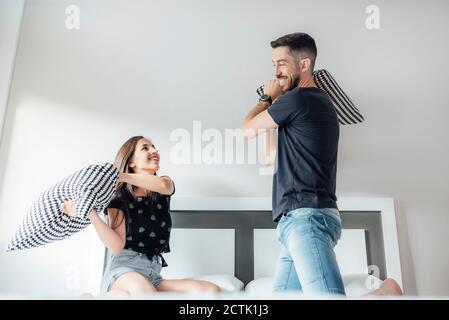 Couple heureux ayant un combat d'oreiller sur le lit dans la chambre à accueil Banque D'Images