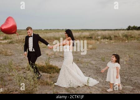 Mariée et marié avec ballon en forme de coeur tenant les mains pendant fille marchant dans le champ Banque D'Images