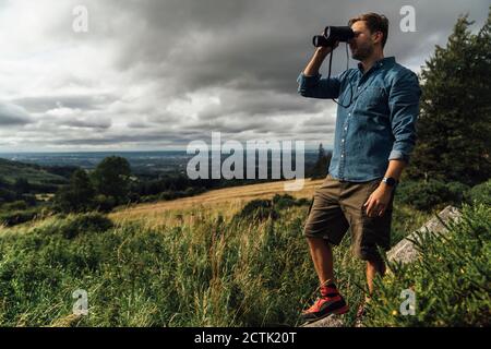 Randonneur mâle regardant à travers des jumelles tout en se tenant sur le paysage contre ciel nuageux Banque D'Images