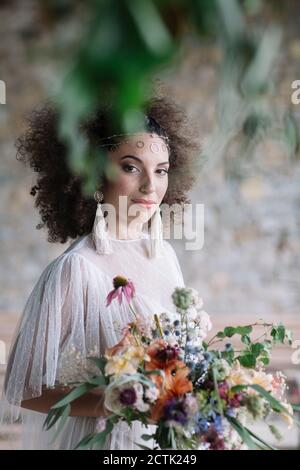 Jeune femme en robe de mariage tenant le bouquet tout en se tenant contre mur Banque D'Images