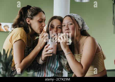 Les adolescentes qui se tiennent dans la cuisine partagent leur smoothie aux fruits frais pailles à boire Banque D'Images