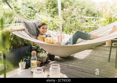 Femme souriante tenant un verre et un smartphone tout en étant couché hamac dans la cour Banque D'Images