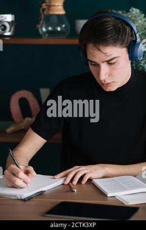 Beau jeune étudiant écrivant dans le livre tout en écoutant de la musique à travers des écouteurs à la maison Banque D'Images