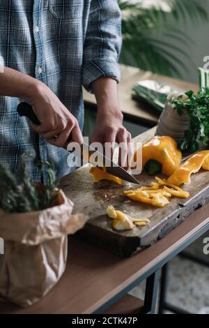 Section médiane du jeune homme coupant du poivron jaune à bord dans la cuisine Banque D'Images